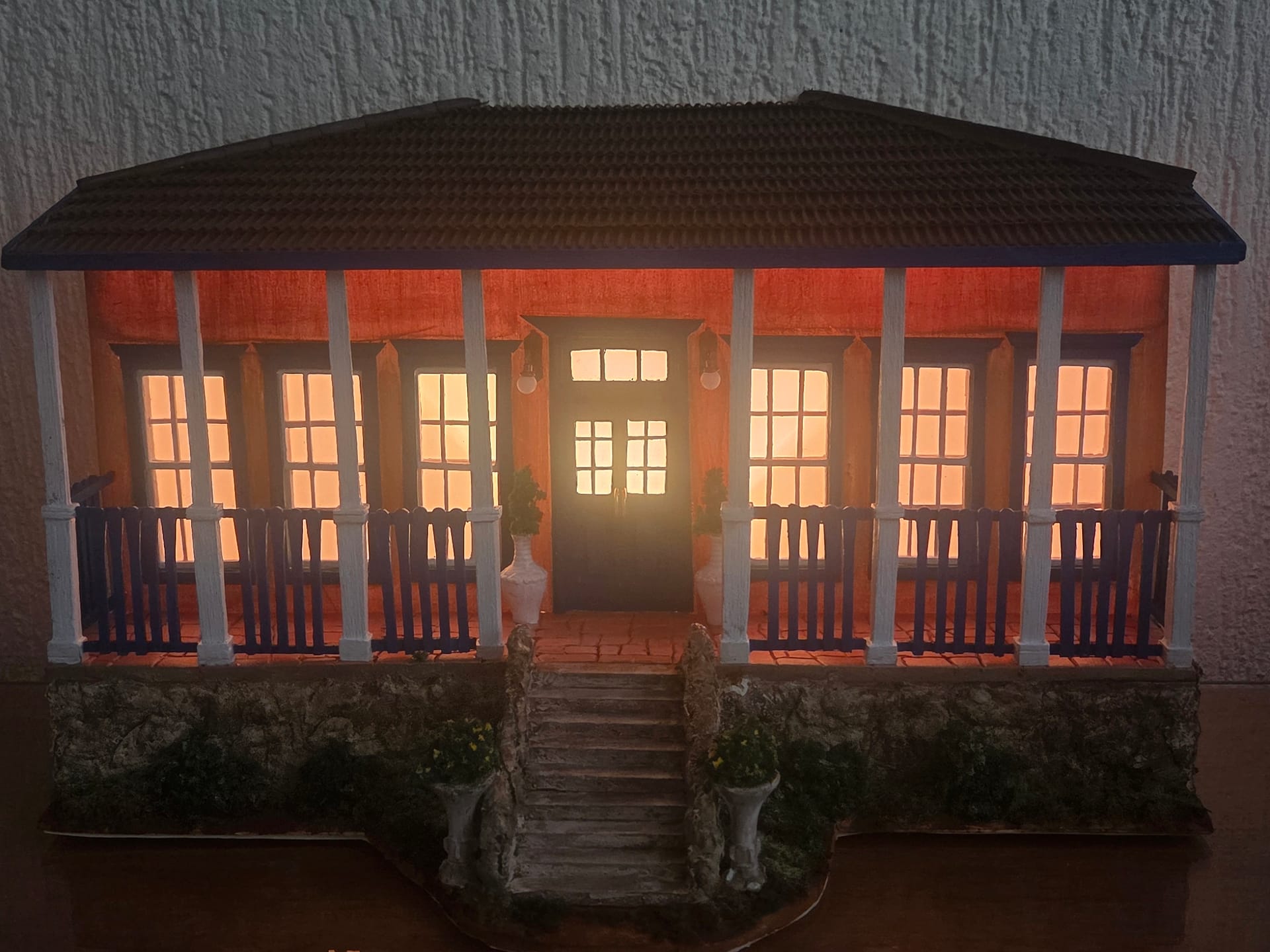 Réplica em miniatura de casa de fazenda com varanda de colunas brancas e janelas iluminadas por dentro, em luz baixa.