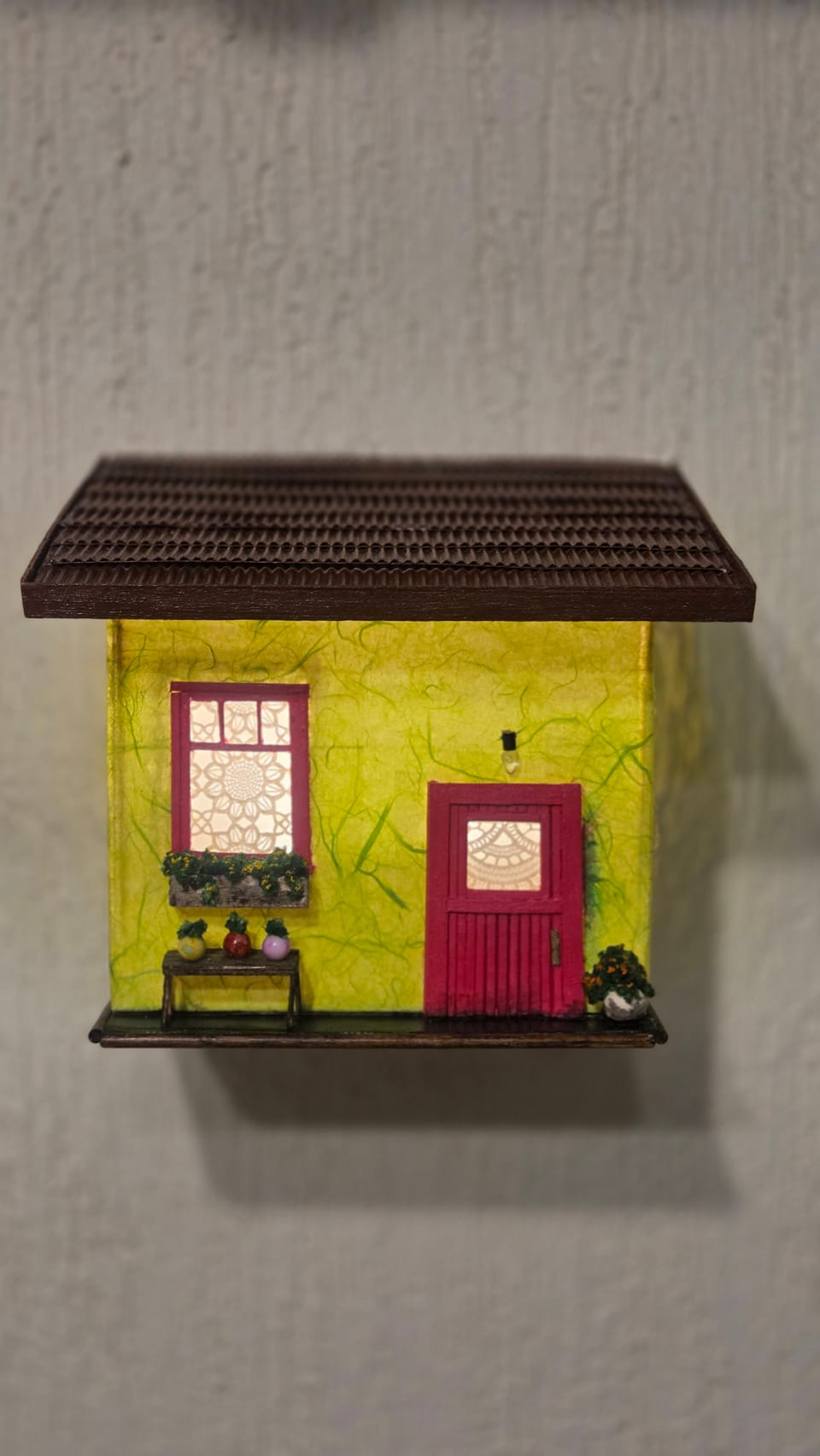 Arandela de parede em forma de casinha amarela com porta e janela rosa, banquinho com vasos, acesa — vista frontal