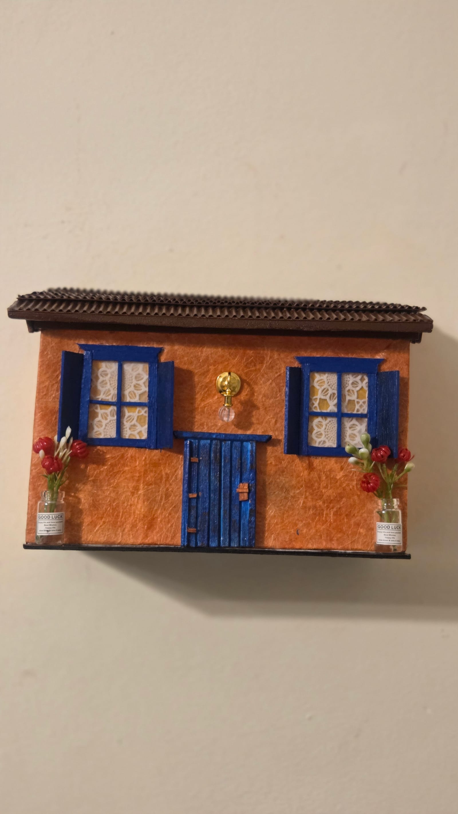 Arandela de parede em forma de casinha laranja com janelas e porta azuis e vasinhos de flores, acesa — vista frontal