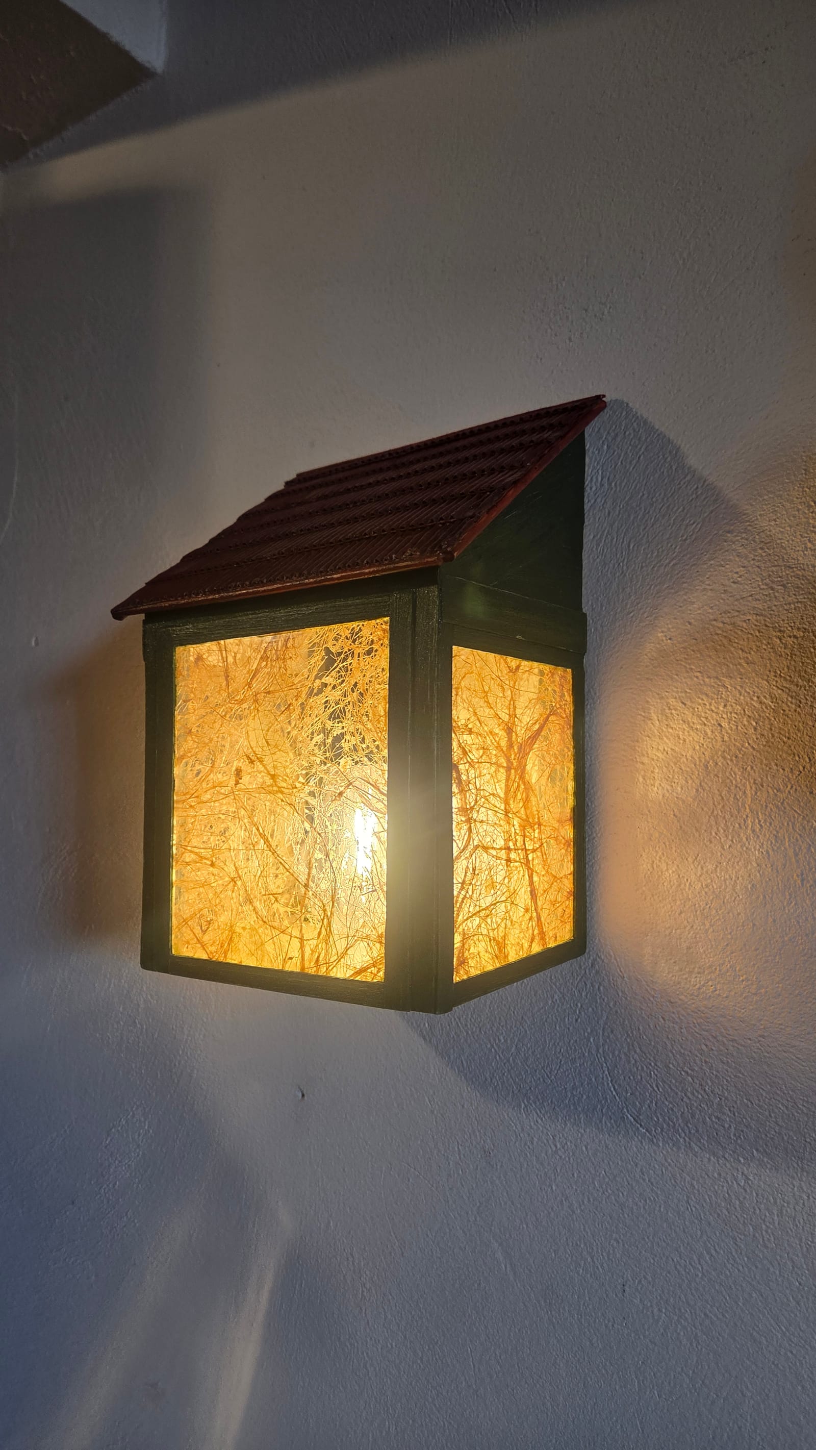 Arandela de parede em forma de lanterna-casinha com papel de fibras naturais iluminado e telhado — acesa, vista em ângulo