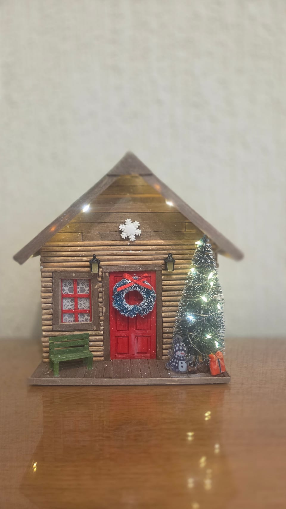 Cabana de madeira de Natal com luzinhas acesas, porta vermelha com guirlanda, árvore e banquinho — vista frontal com as luzes acesas