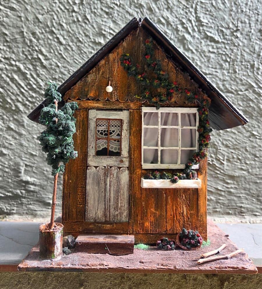 Cabana rústica de madeira em miniatura com porta branca envelhecida, janela com cortina, árvore e telhado escuro — vista frontal