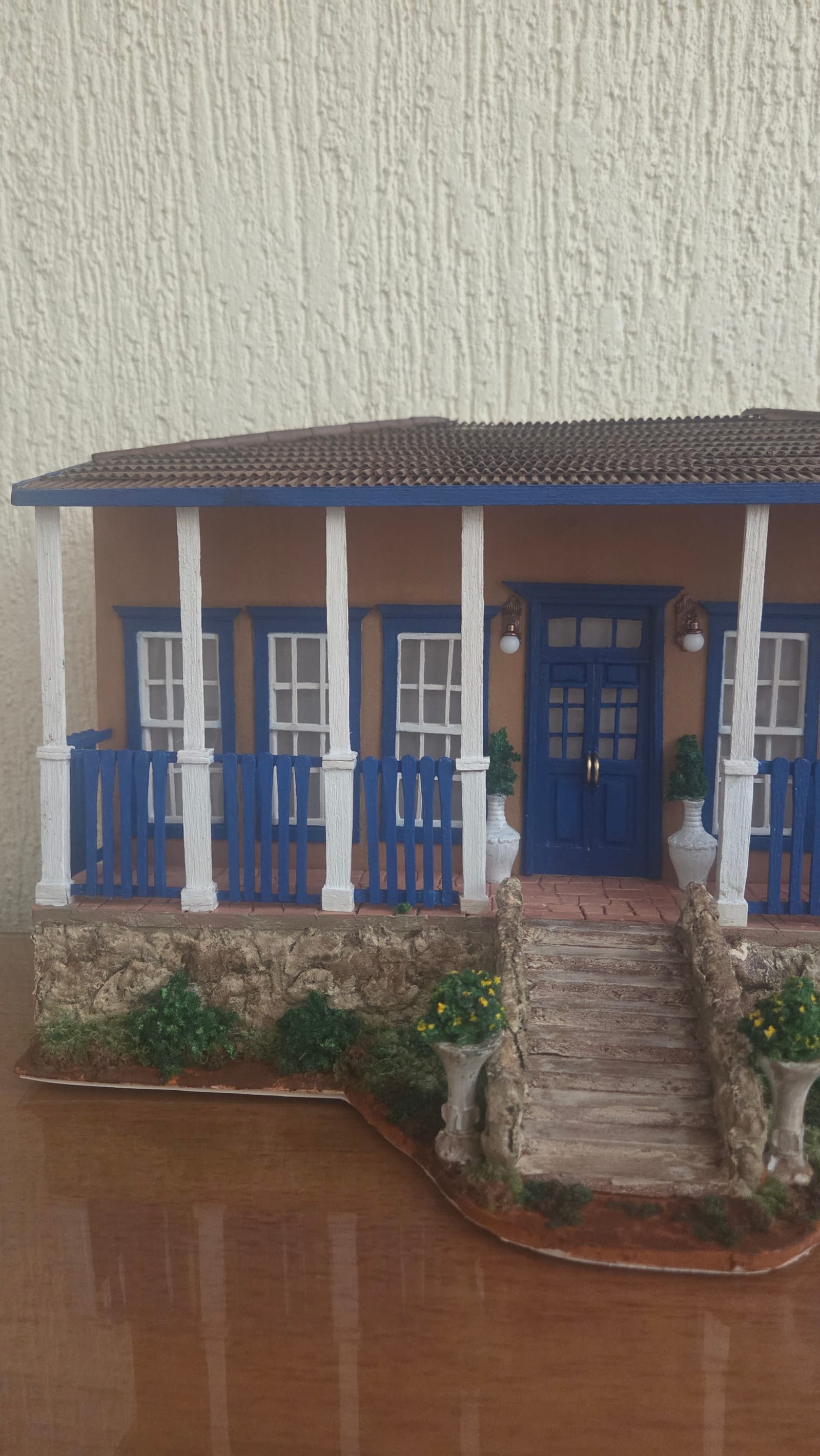 Casa de fazenda em miniatura com fachada ocre, portas e gradis azuis, colunas brancas, varanda e escadaria de pedra — vista frontal