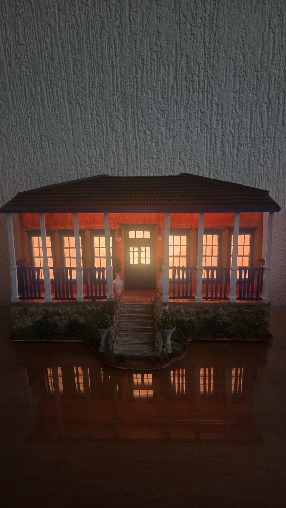 Casa de fazenda em miniatura com fachada ocre, portas e gradis azuis, colunas brancas, varanda e escadaria de pedra — com a iluminação acesa