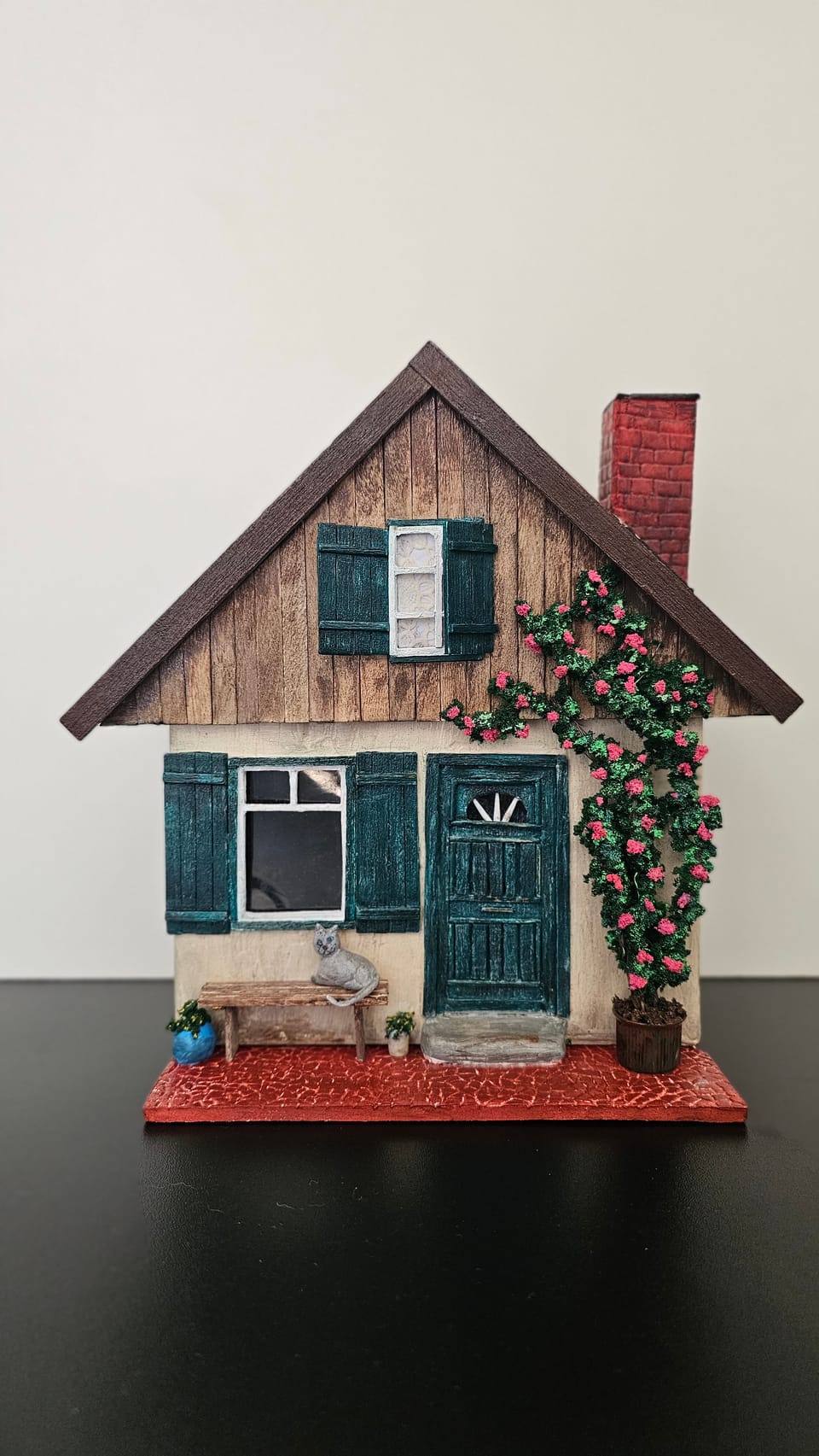 Casinha em miniatura com roseira na fachada, janelas verdes, chaminé de tijolos, banco e gatinho na varanda — vista frontal