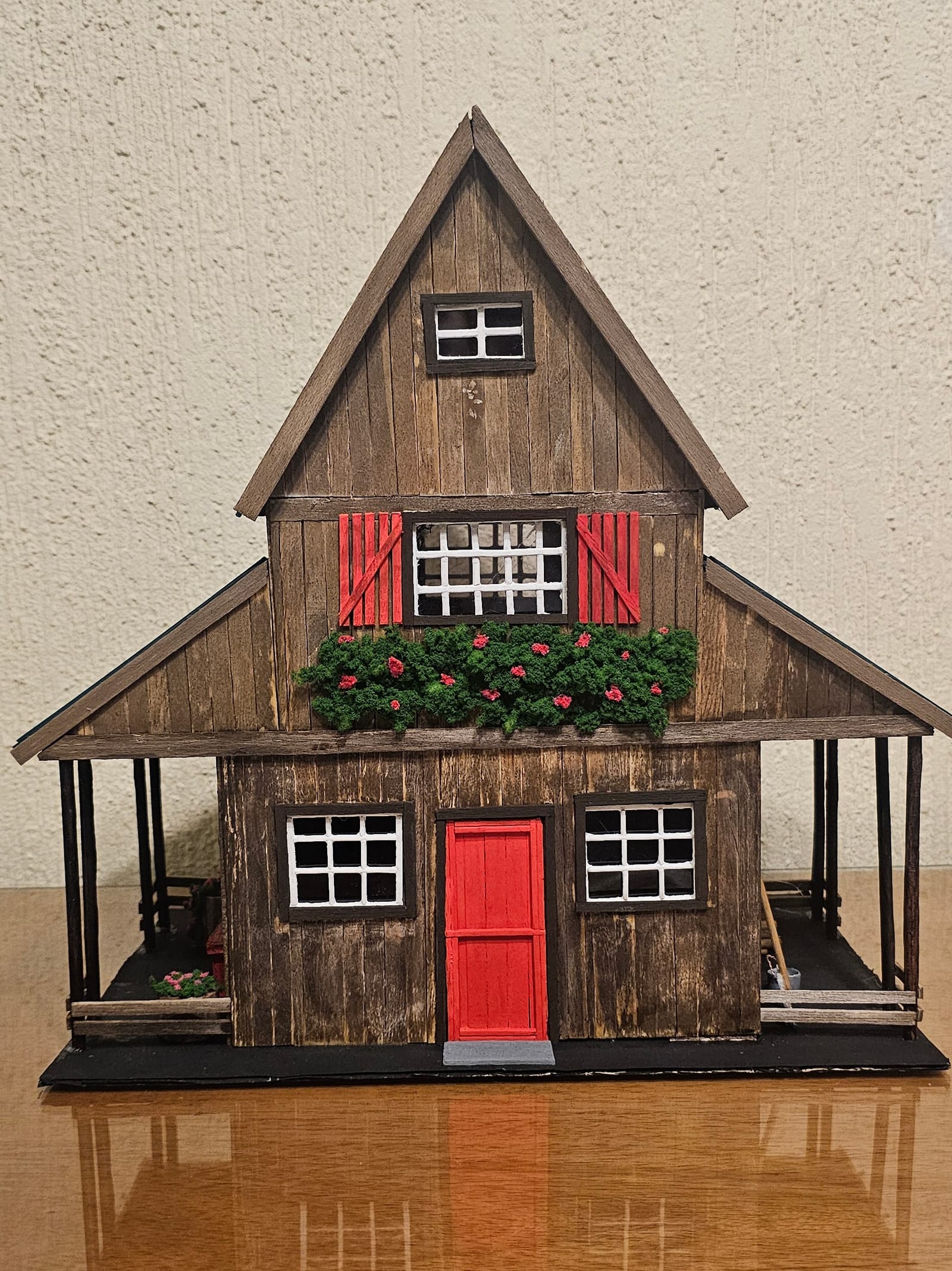 Casa de madeira em miniatura com telhado verde-azulado, porta e venezianas vermelhas, floreira e varanda — vista frontal