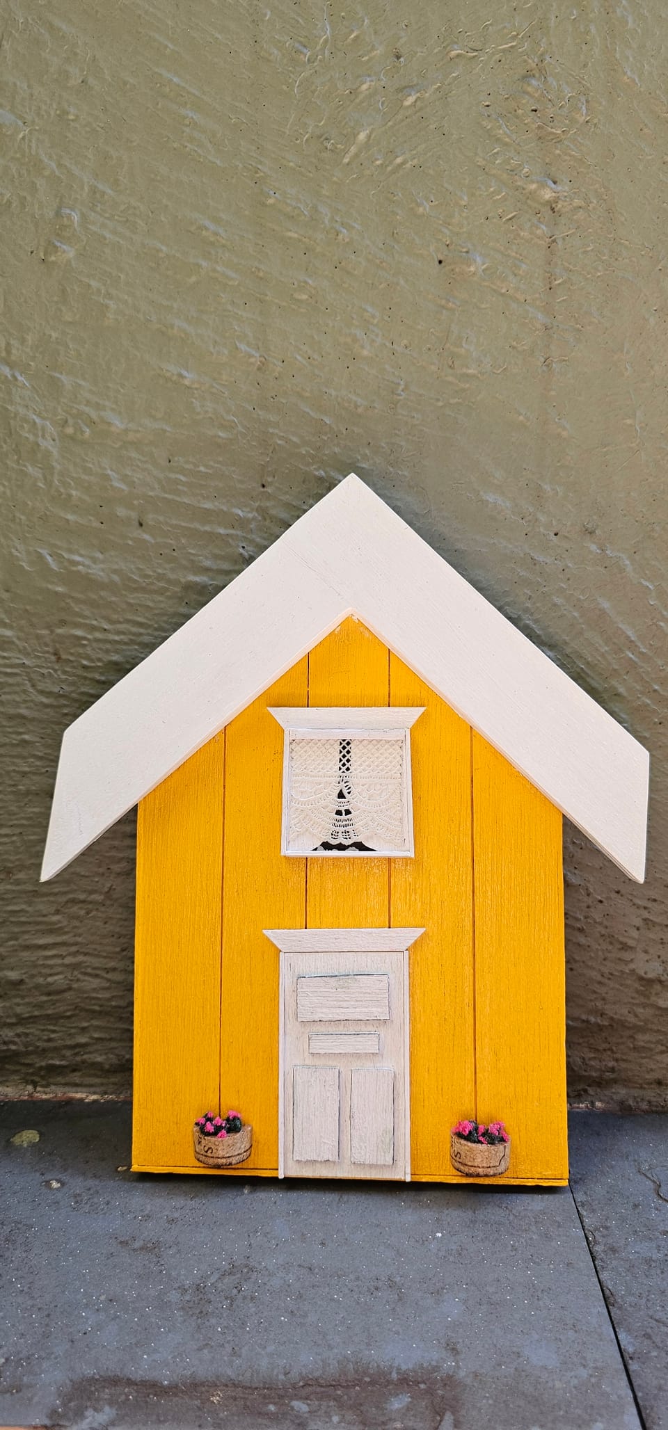Fachada de casinha amarela de tábuas com telhado branco, janela com cortina de renda, porta branca e vasinhos de flores — vista frontal