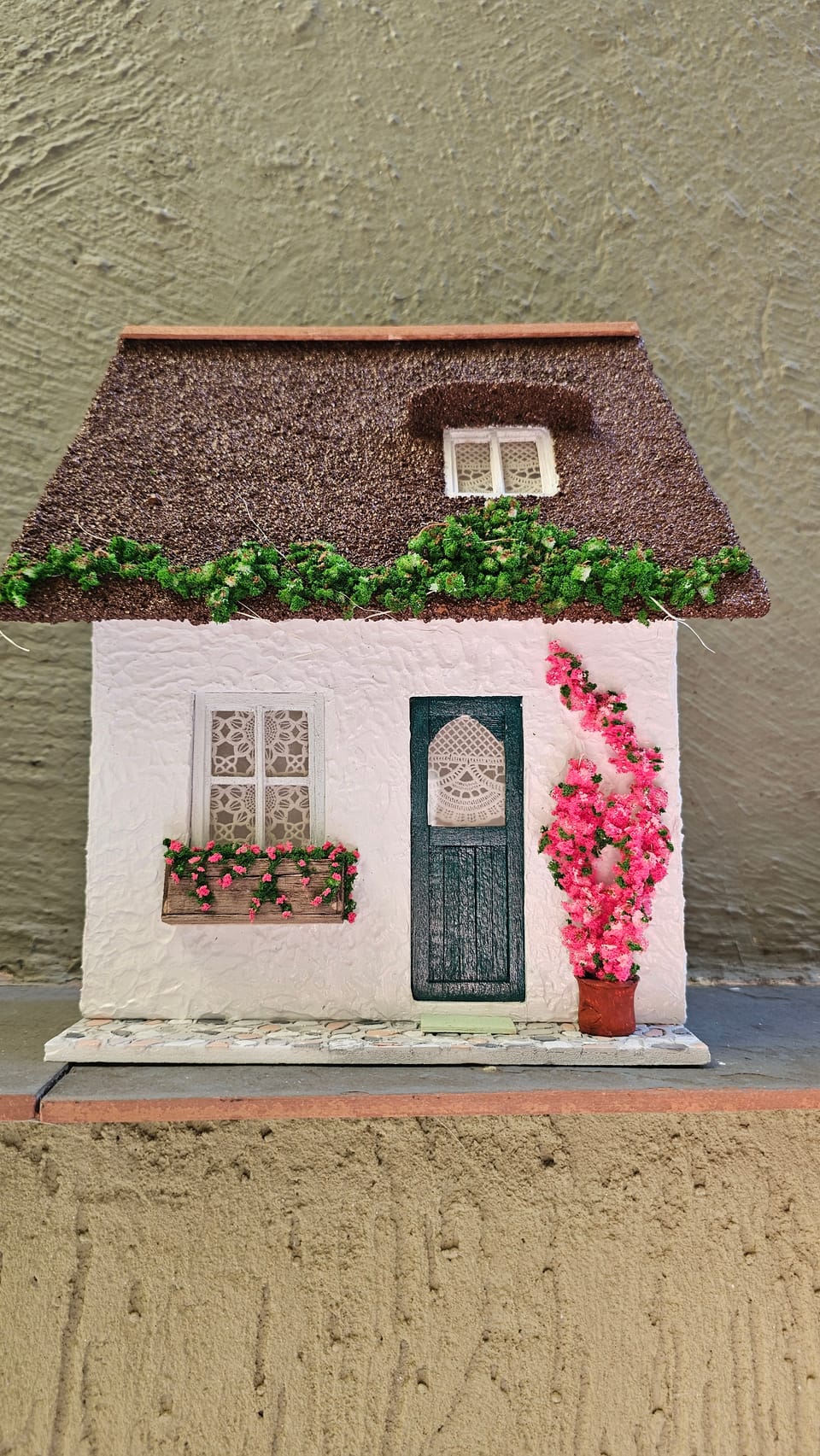 Chalé branco em miniatura com telhado de palha marrom, trepadeira, porta verde e flores rosa — vista frontal