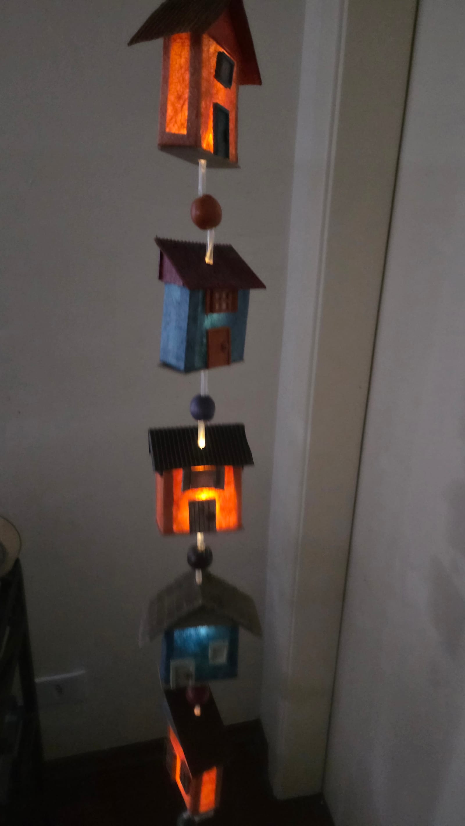 Cordão vertical de casinhas penduradas com luz acesa por dentro, intercaladas com contas de madeira — detalhe das casinhas acesas