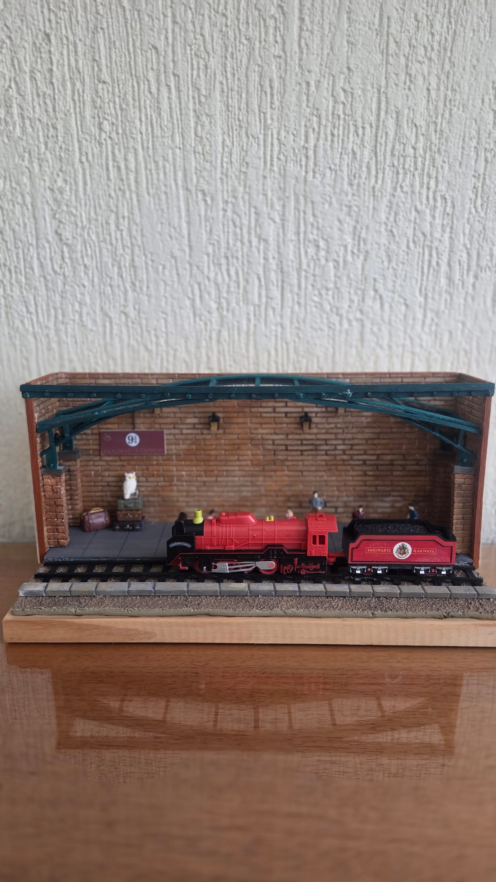 Diorama de estação de trem com parede de tijolinhos, cobertura de ferro verde, locomotiva vermelha, malas e gaiola de coruja — vista frontal