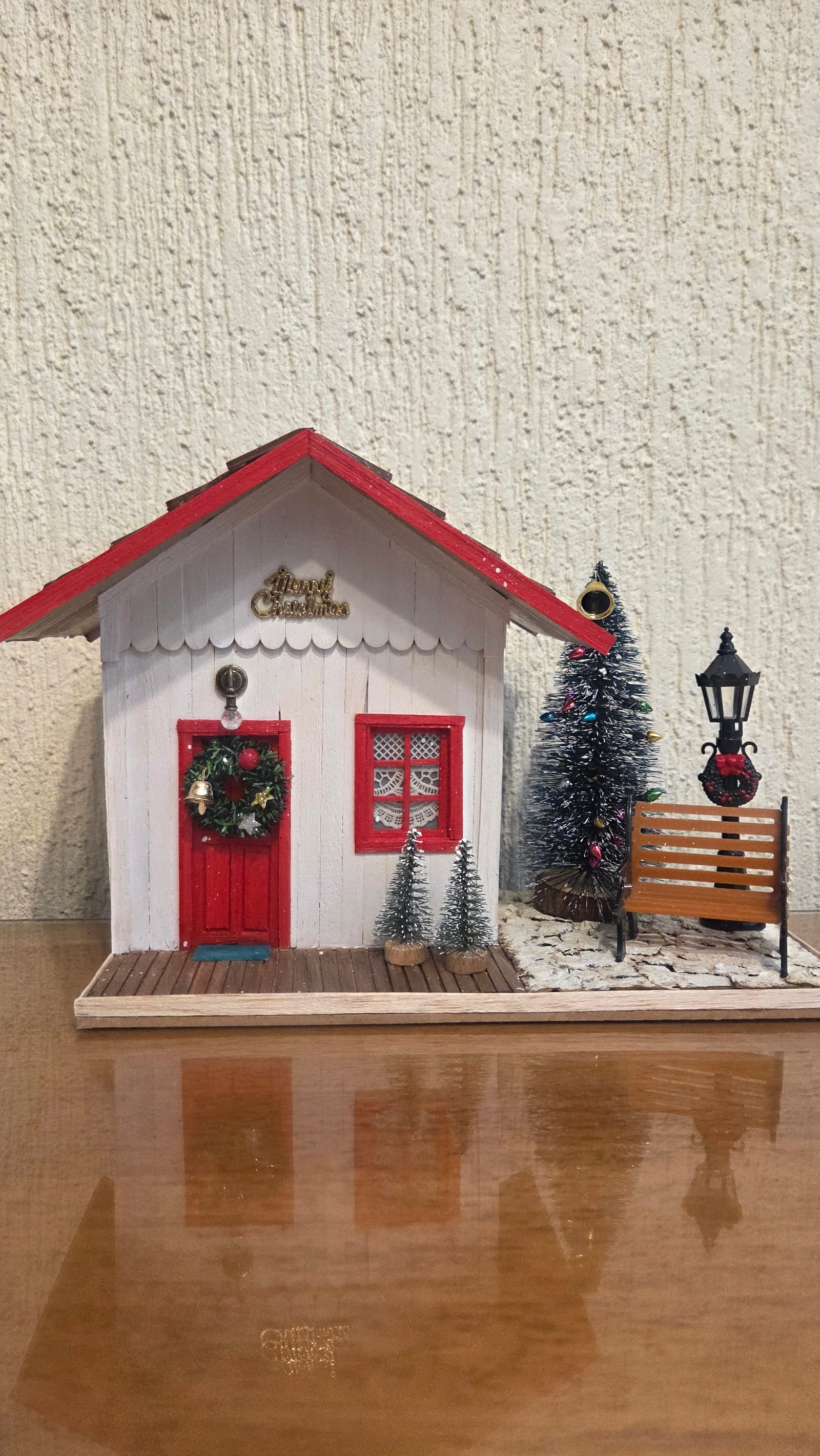Diorama de Natal de uma casinha branca de telhado vermelho, porta com guirlanda, árvore, poste e banco — vista frontal