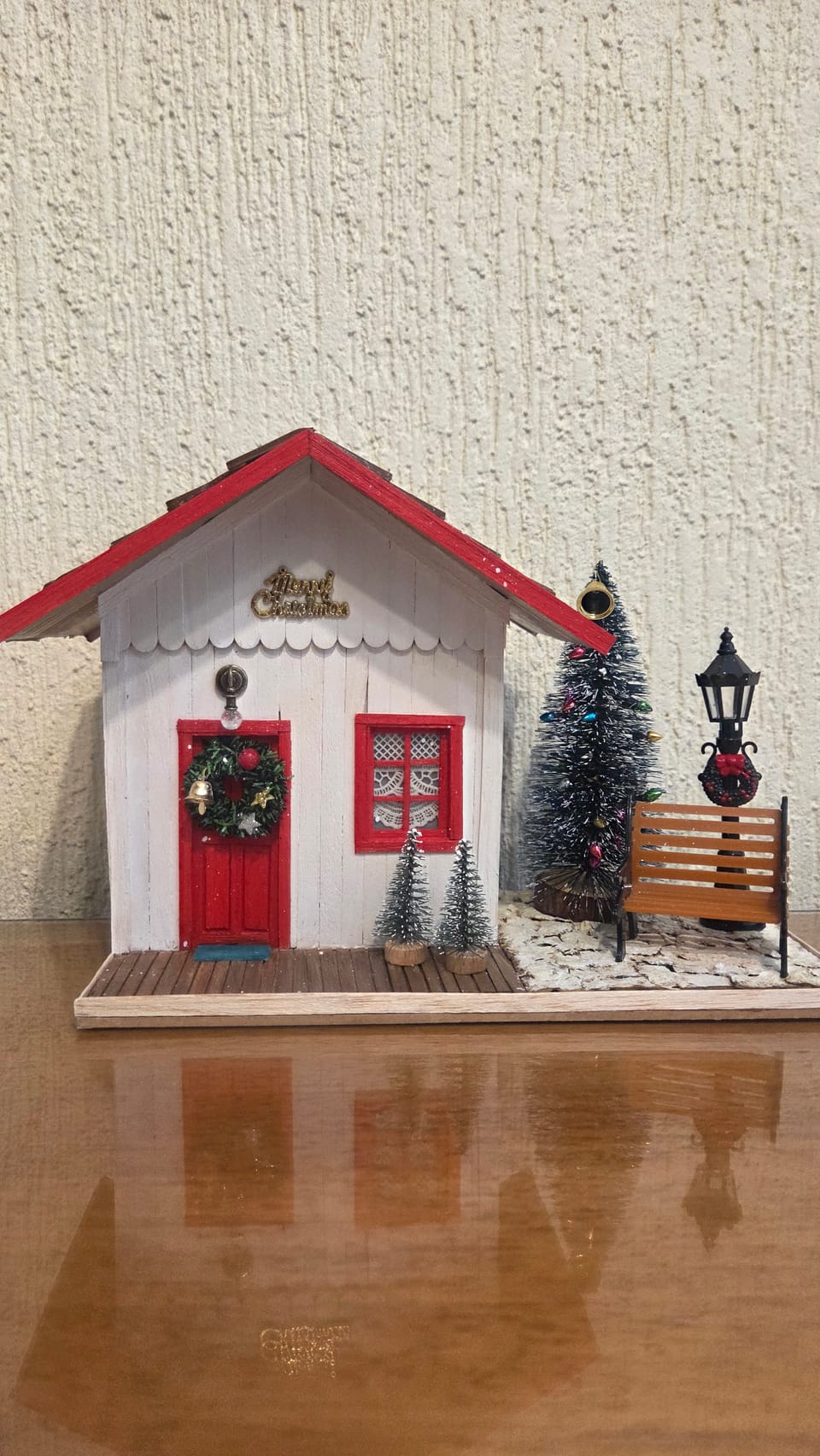 Diorama de Natal de uma casinha branca de telhado vermelho, porta com guirlanda, árvore, poste e banco — vista frontal
