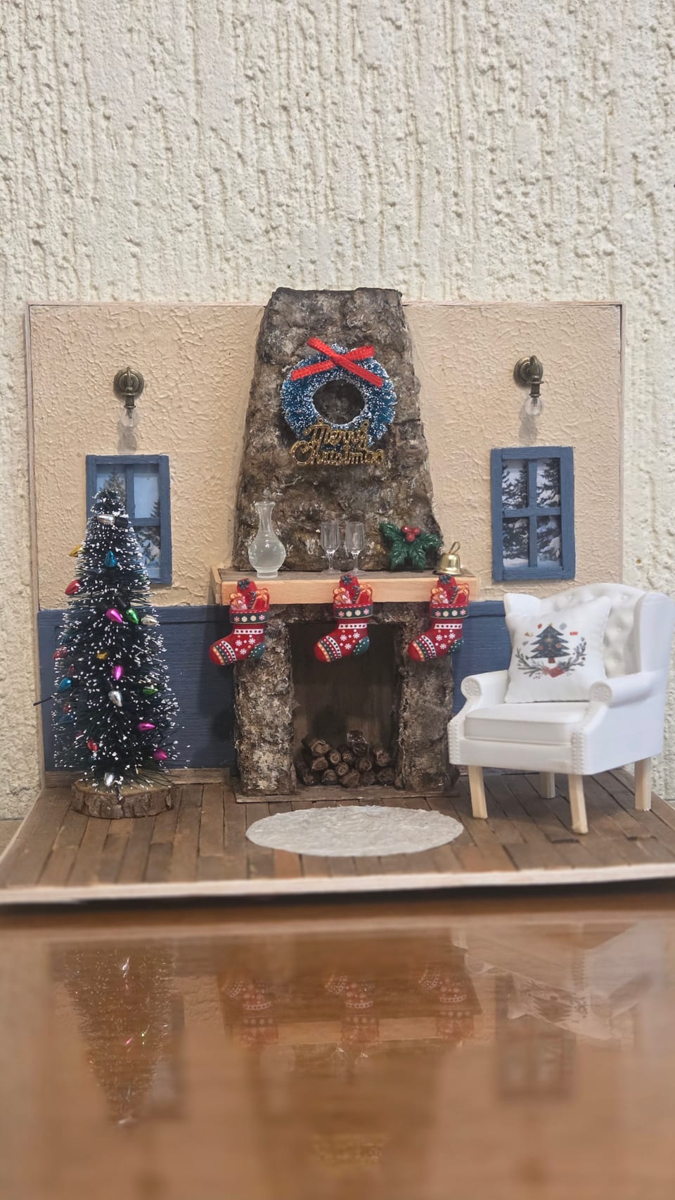 Diorama de Natal de uma sala com lareira de pedra, guirlanda, meias, árvore com luzinhas e poltrona branca — vista frontal