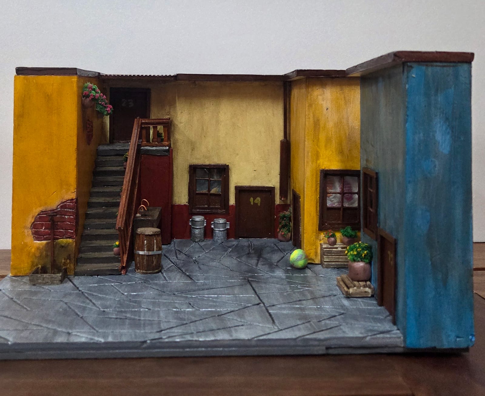 Diorama de vila com paredes amarelas, escadaria de madeira, barril, porta número 14 e pátio de pedras — vista frontal