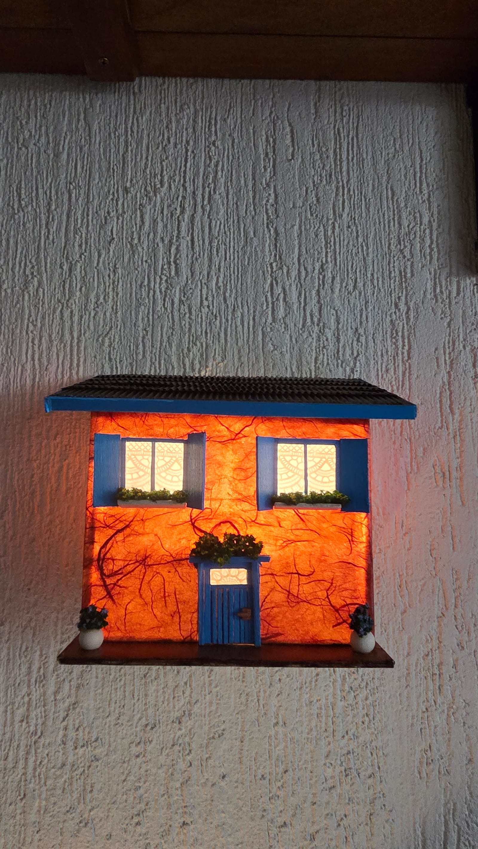 Luminária de parede em forma de sobrado laranja com janelas e porta azuis, acesa — acesa