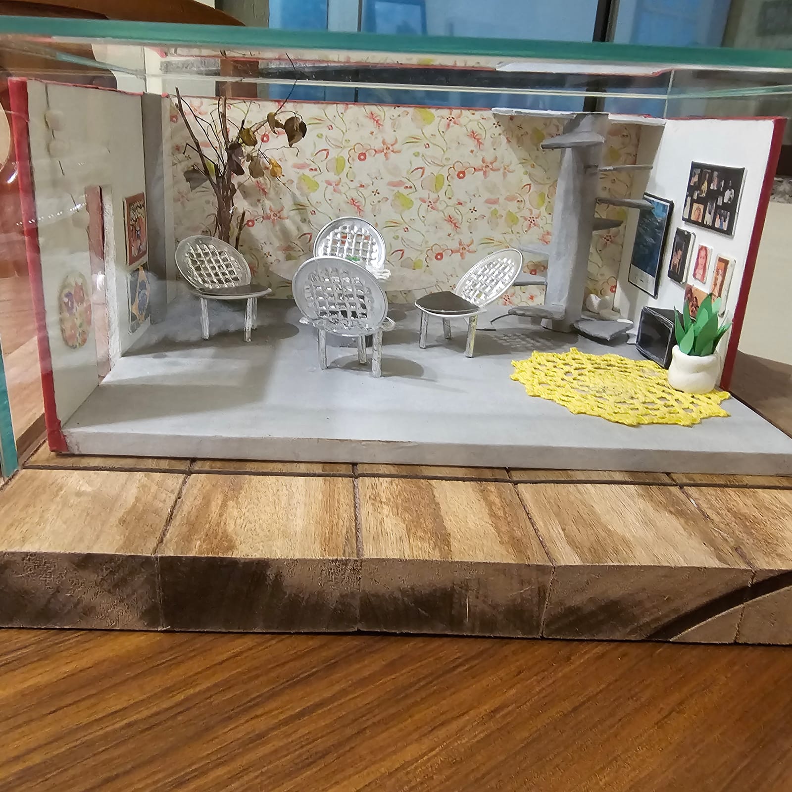 Mini ambiente com papel de parede florido, cadeiras de fio, toalhinha de crochê amarela, escada caracol e quadrinhos — vista frontal, na caixa de vidro