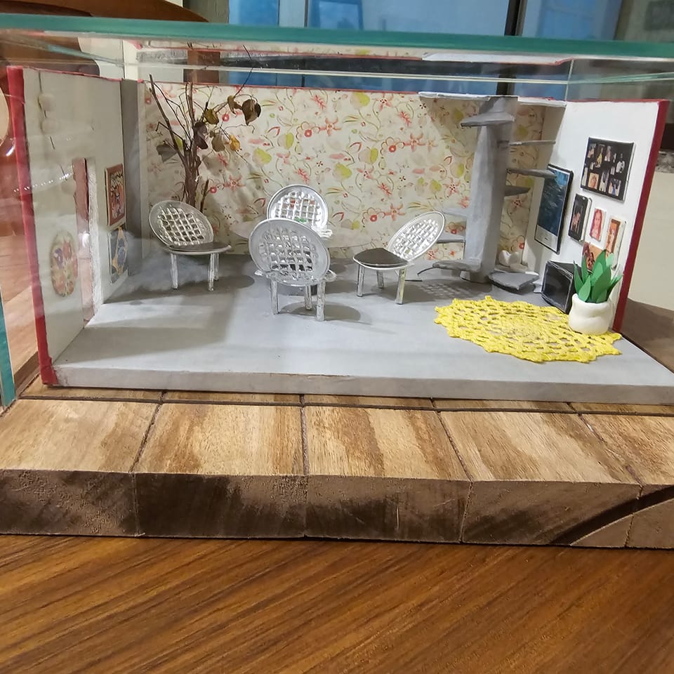 Mini ambiente com papel de parede florido, cadeiras de fio, toalhinha de crochê amarela, escada caracol e quadrinhos — vista frontal, na caixa de vidro