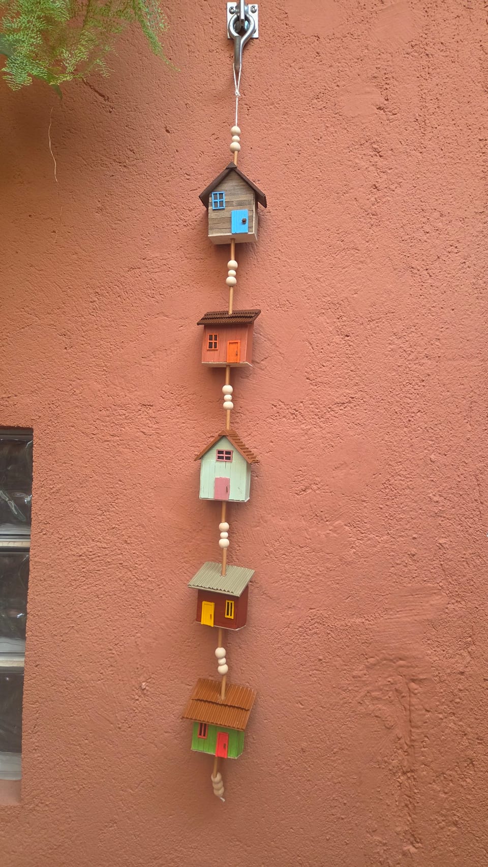 Móbile vertical com cinco casinhas de madeira coloridas intercaladas com contas, pendurado na parede — vista completa, com a casinha azul-acinzentada no alto
