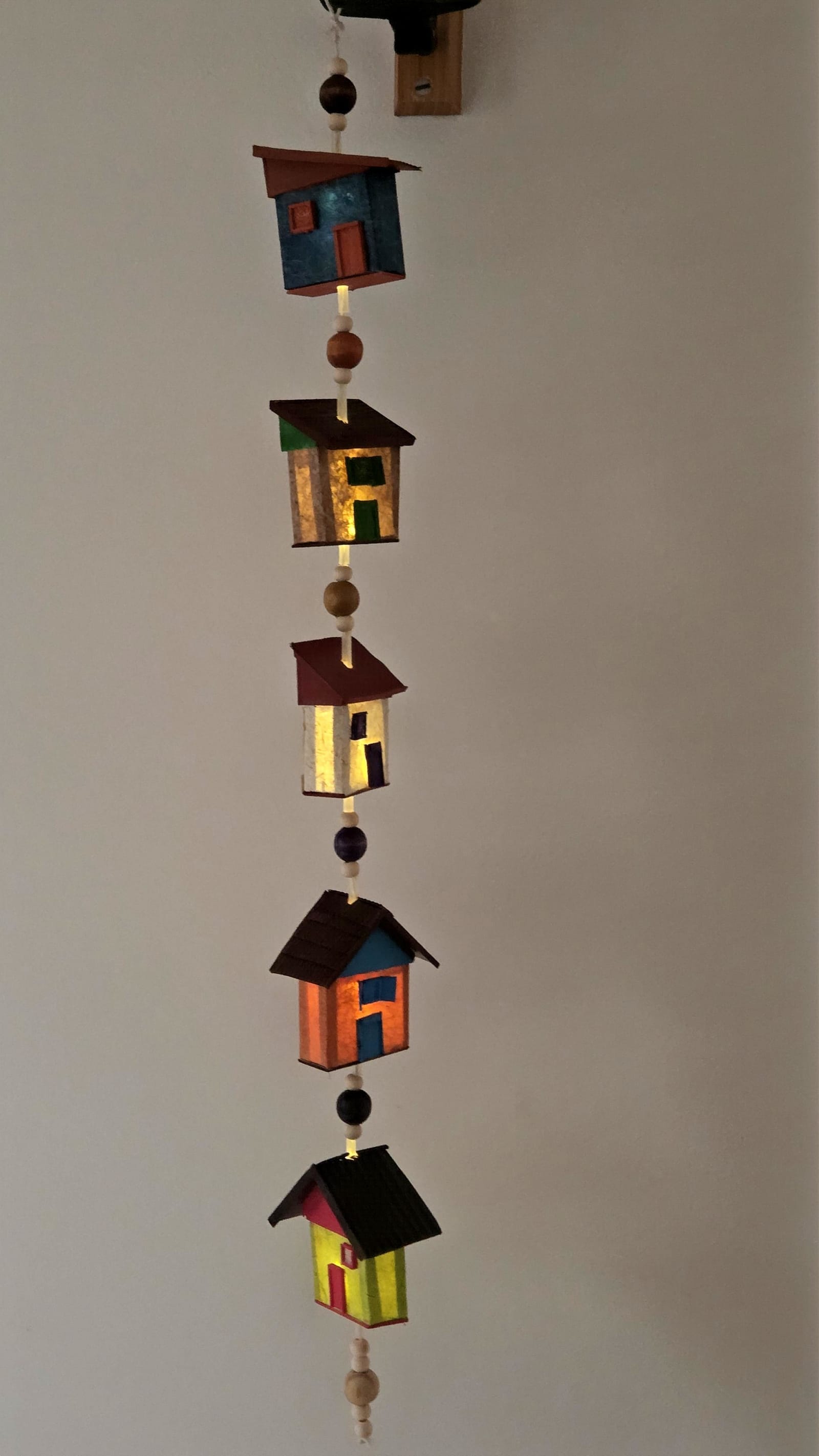 Móbile vertical de casinhas coloridas com luz acesa por dentro, intercaladas com contas — vista frontal