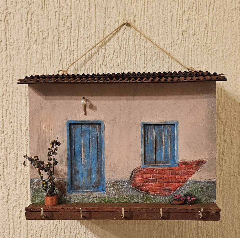 Porta-chaves de parede em forma de fachada rústica com porta e janela azuis, tijolinhos aparentes e vaso de planta — vista frontal