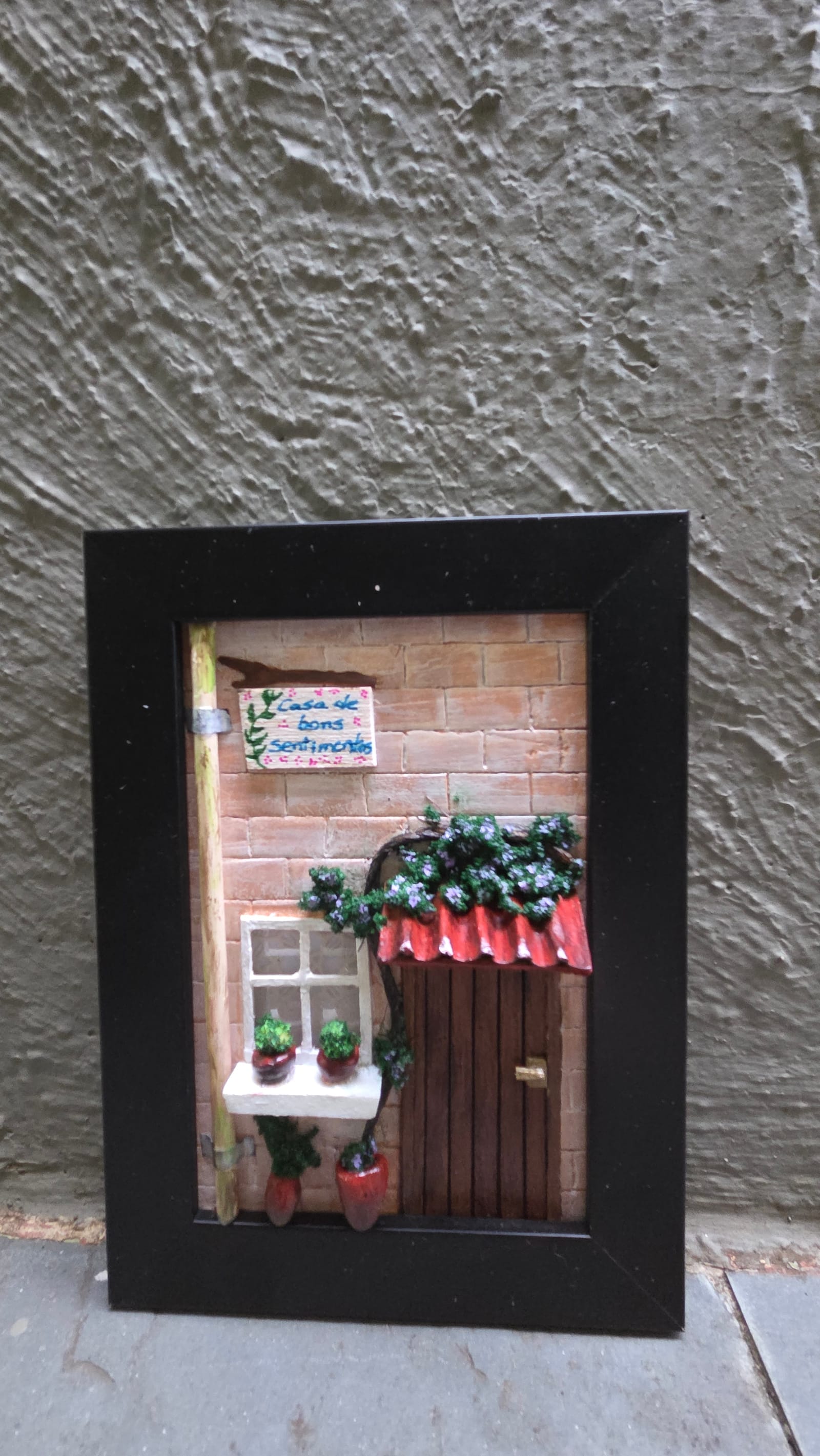 Quadro com fachada de tijolinhos, plaquinha "Casa de bons sentimentos", janela, porta e trepadeira — vista frontal