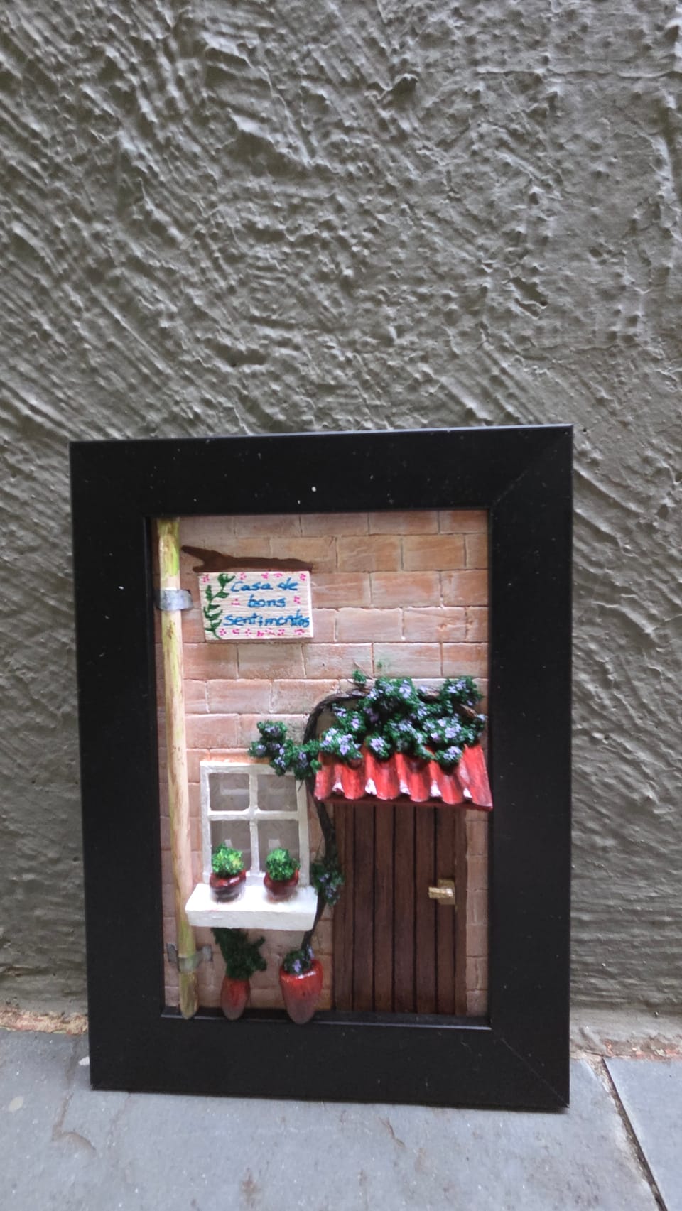 Quadro com fachada de tijolinhos, plaquinha "Casa de bons sentimentos", janela, porta e trepadeira — vista frontal