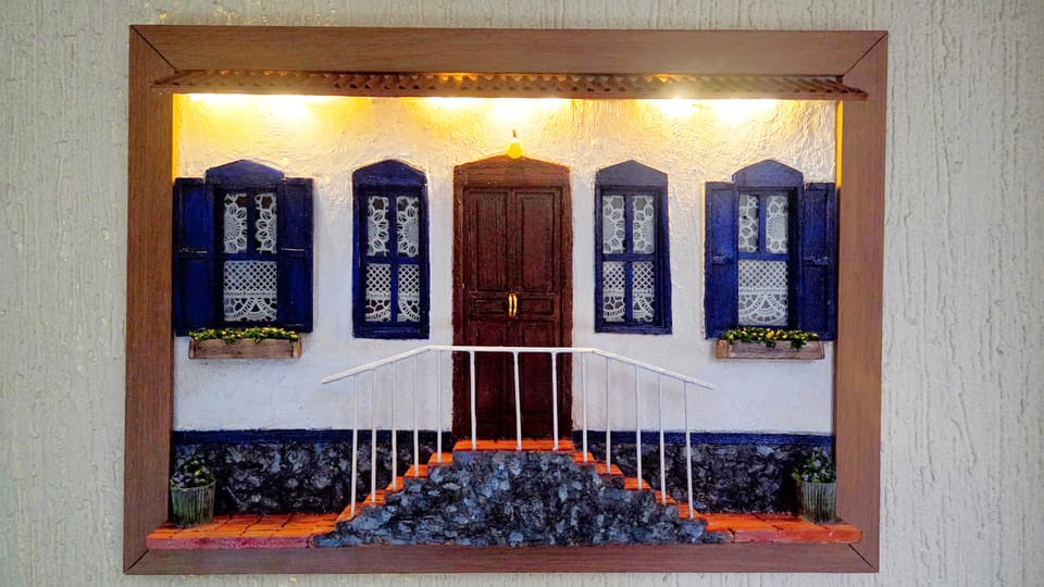 Fachada de casa de fazenda branca com janelas azuis, porta de madeira, escadaria de pedra e luzinhas sob o beiral — com a iluminação acesa