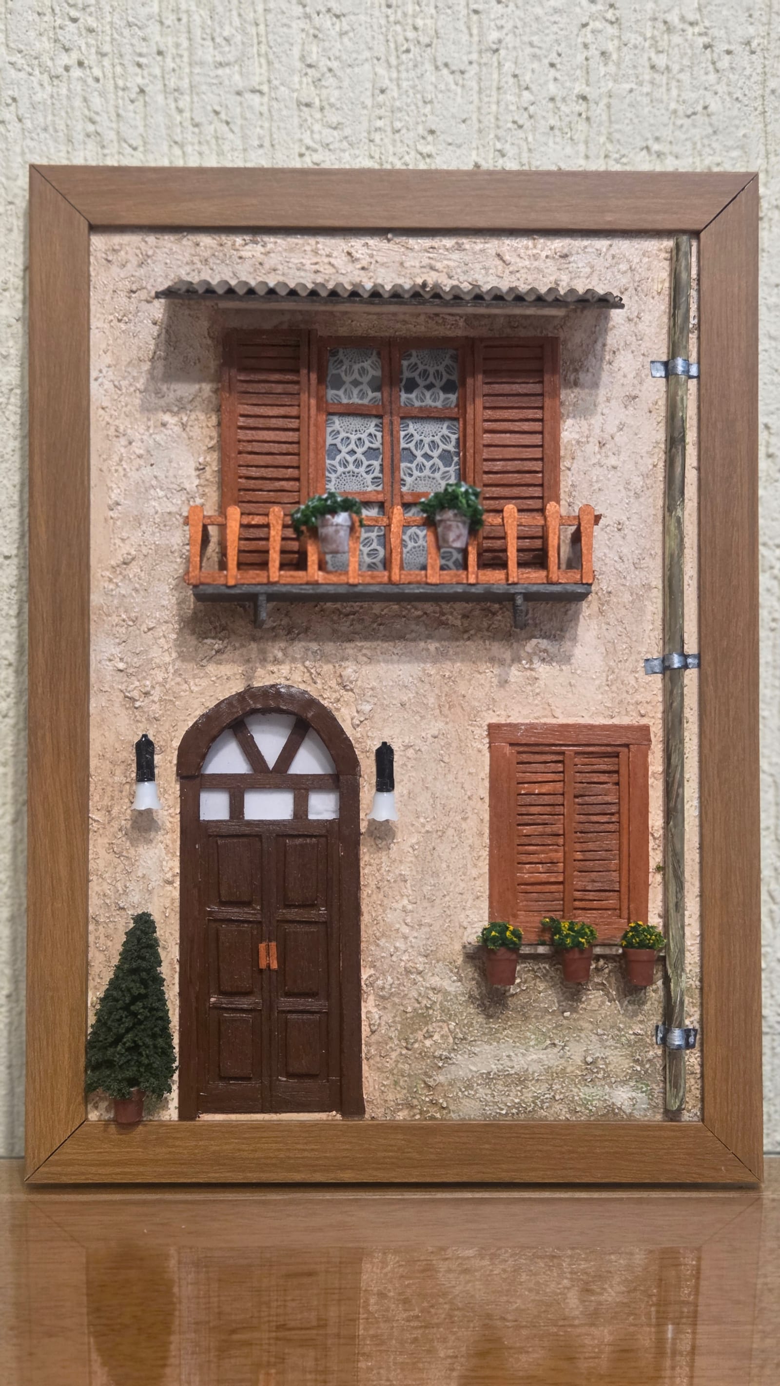 Quadro com fachada rústica em relevo: porta em arco, sacada com vasinhos, janelas de venezianas e arandelas, em moldura de madeira — vista frontal