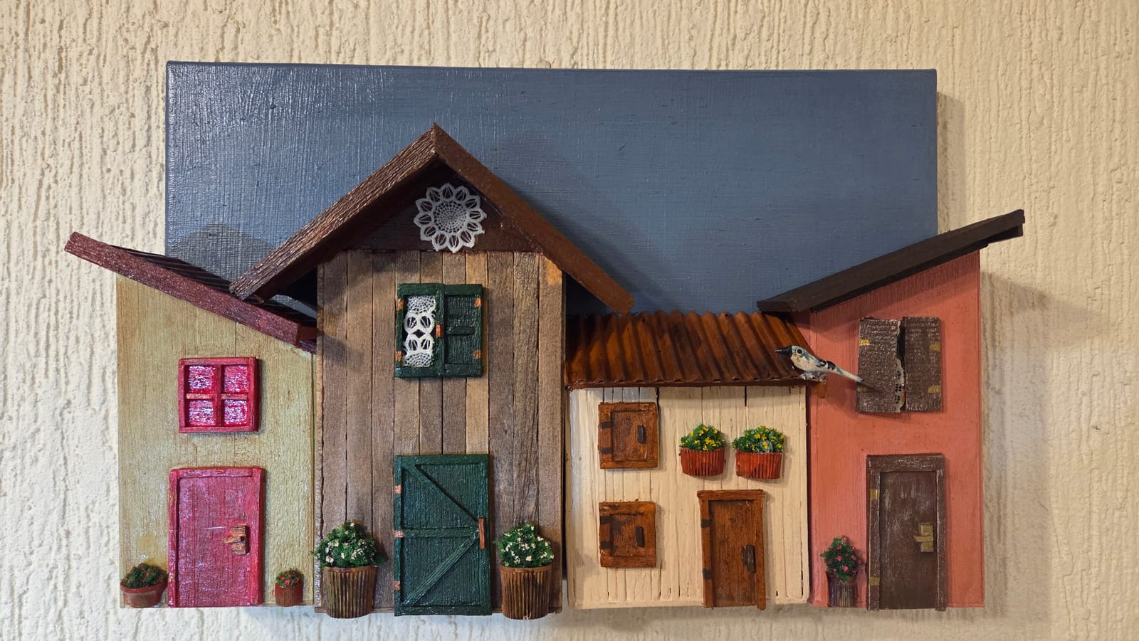 Quadro com duas casinhas de madeira, porta verde, janela rendada, floreiras e fundo azul — vista frontal