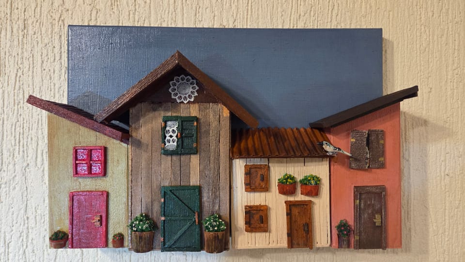 Quadro com duas casinhas de madeira, porta verde, janela rendada, floreiras e fundo azul — vista frontal