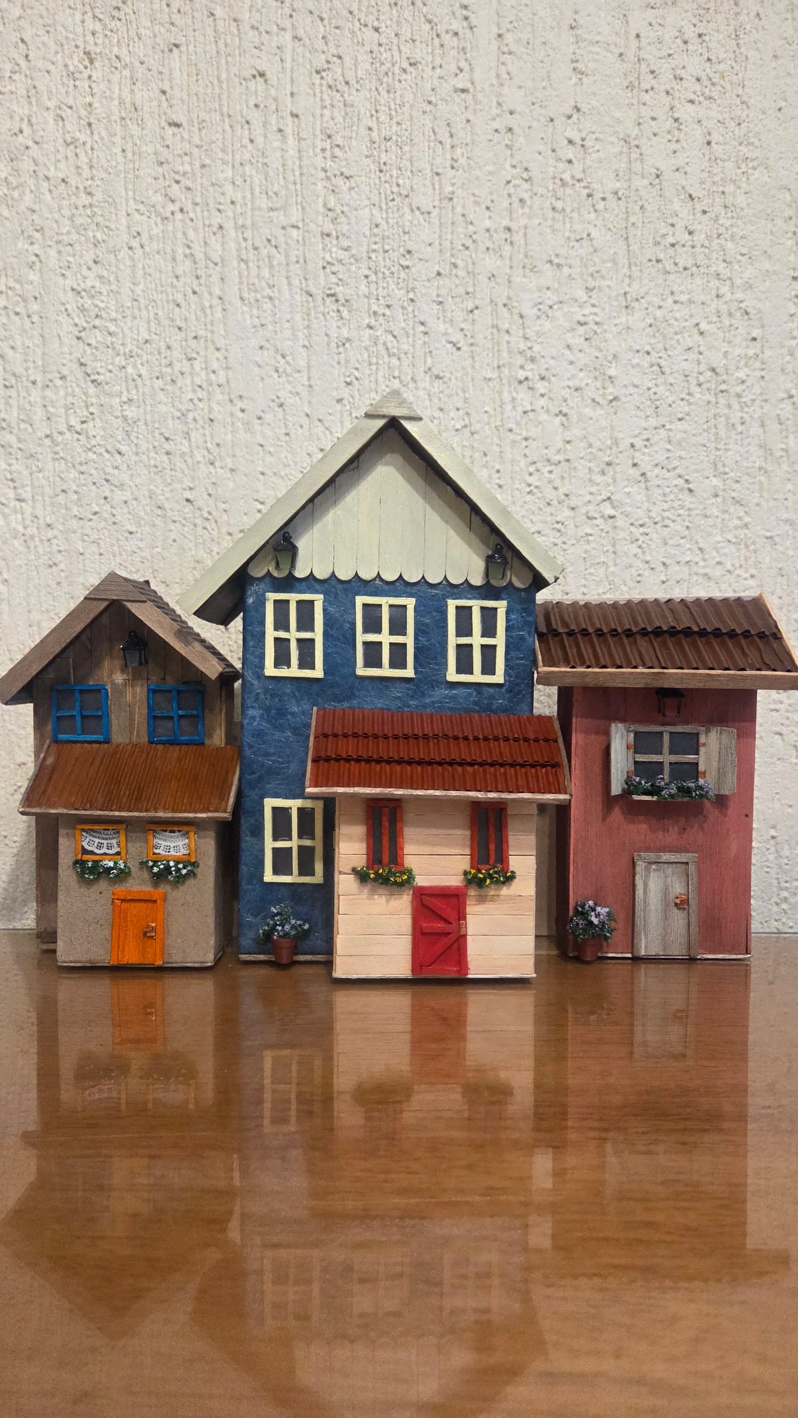 Vila de casinhas em miniatura lado a lado: sobrado azul, casinha creme de porta vermelha, casinha de janelas azuis e casinha rosa — vista frontal