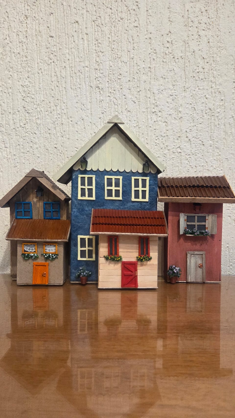 Vila de casinhas em miniatura lado a lado: sobrado azul, casinha creme de porta vermelha, casinha de janelas azuis e casinha rosa — vista frontal