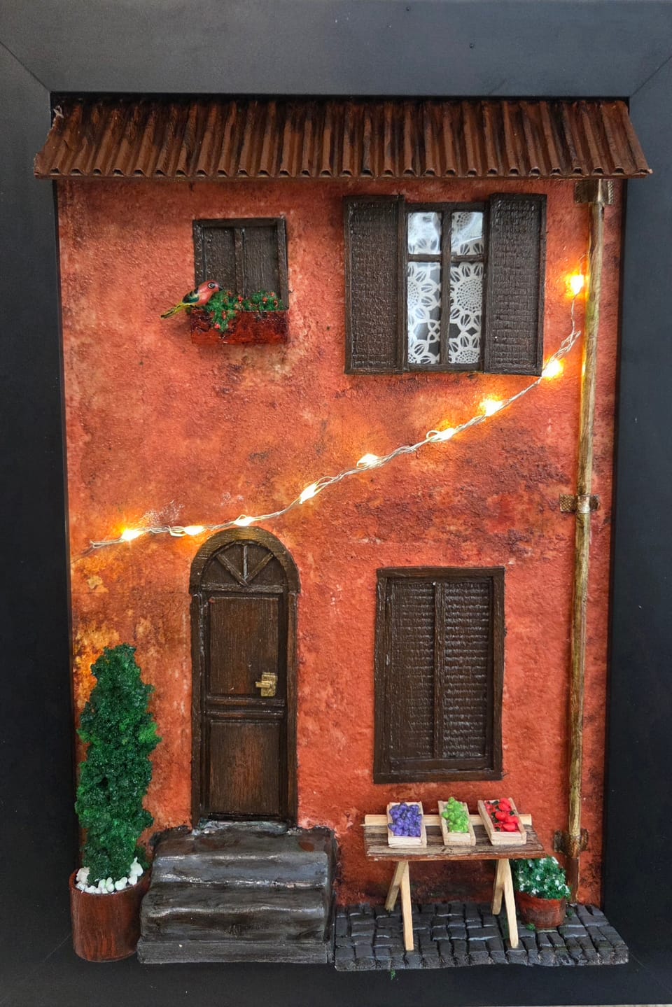 Quadro com fachada italiana alaranjada, cordão de luzinhas, banca de frutas e passarinho na janela — vista frontal