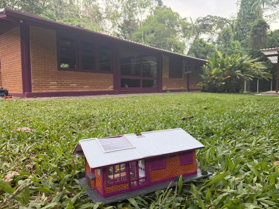 Réplica de casa térrea de tijolinhos com detalhes em vinho, telhado claro removível e interior mobiliado — diante da casa original, no jardim